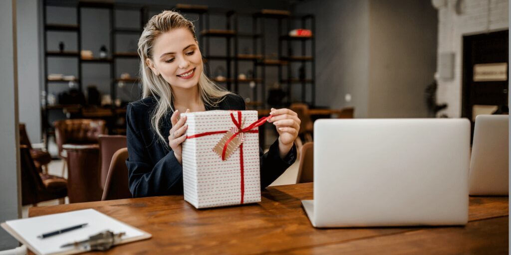 A woman sits at a wooden desk in an office, smiling while unwrapping a gift box. A laptop, clipboard, and other items are on the desk.
