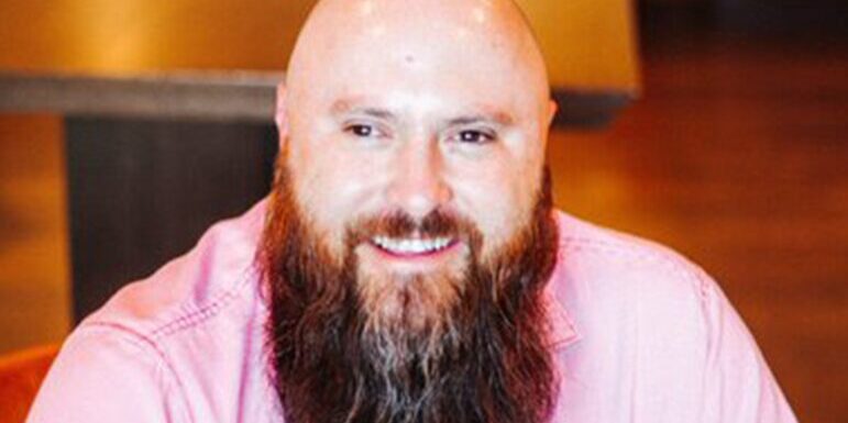 A bearded man smiles at the camera with blurred background.