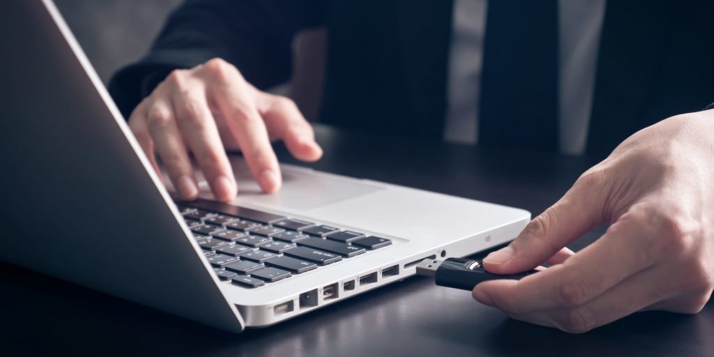 A person inserts a USB flash drive into a laptop on a dark table, with one hand on the keyboard and the other holding the flash drive.