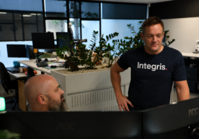 Two men work in a modern office with plants and computer monitors; one stands wearing an “Integris.” shirt, while the other sits at a desk—collaborating on projects powered by CORE Managed AI and IT Services.