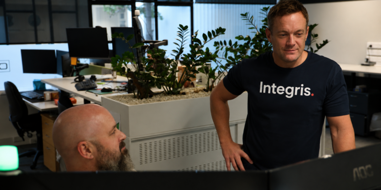 Two men work in a modern office with plants and computer monitors; one stands wearing an “Integris.” shirt, while the other sits at a desk—collaborating on projects powered by CORE Managed AI and IT Services.