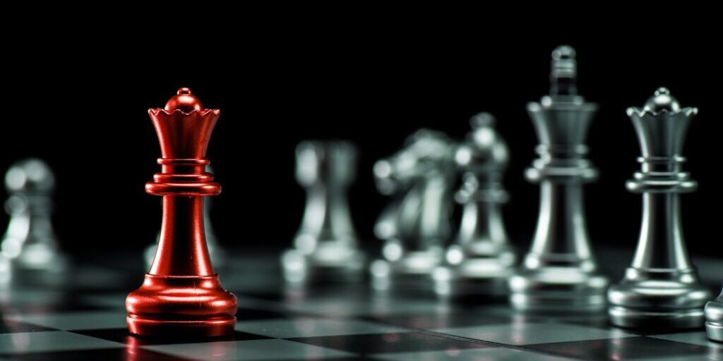 A red queen chess piece is prominent in the foreground on a chessboard, contrasting with silver chess pieces blurred in the background on a black backdrop. The image highlights the uniqueness and significance of the red queen amid the standard silver pieces.
