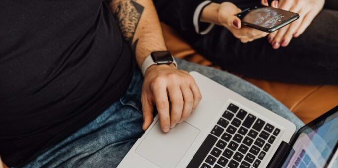 Two people sitting on a couch; one is engrossed in a laptop, while the other is captivated by a smartphone.