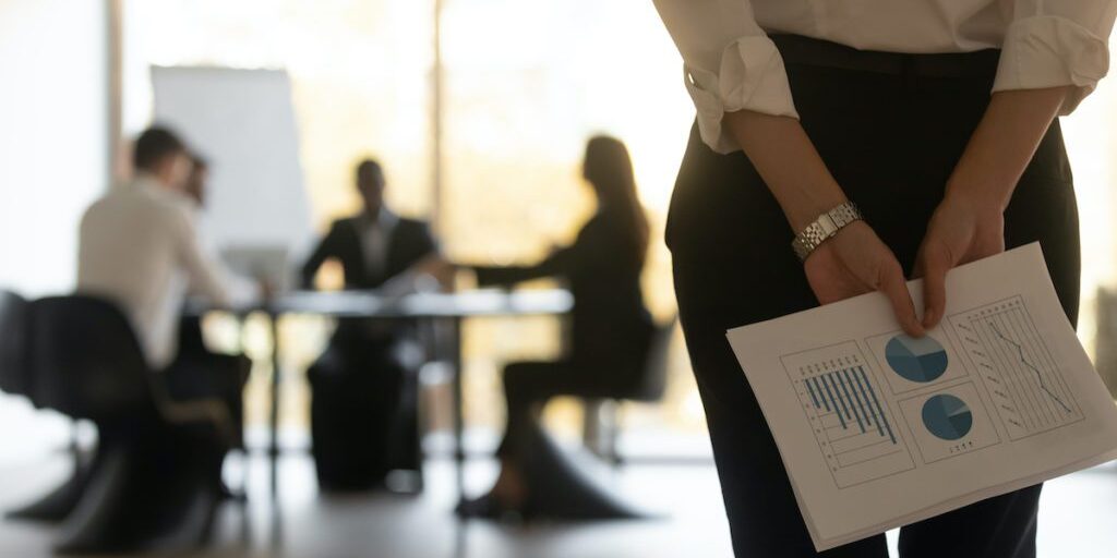 A person holding printed charts stands in the foreground while four individuals sit at a table in a business meeting background.