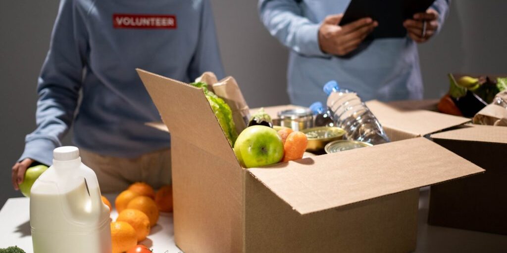 Two people are sorting food items into cardboard boxes. One is holding a green apple while various groceries, including milk, tomatoes, and water bottles, are on the table. Another person is holding a clipboard and meticulously checking off items as they go.