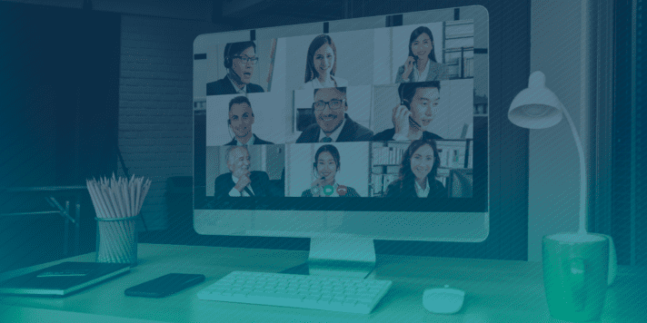 A computer screen displays a video conference with nine participants. A desk lamp casts a soft glow, and a mug and stationery are neatly arranged on the desk in front of the monitor.