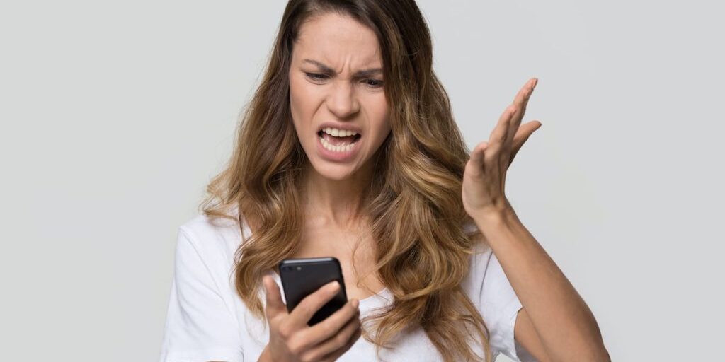 A woman in a white shirt looks at her phone with an angry expression, raising one hand in frustration.