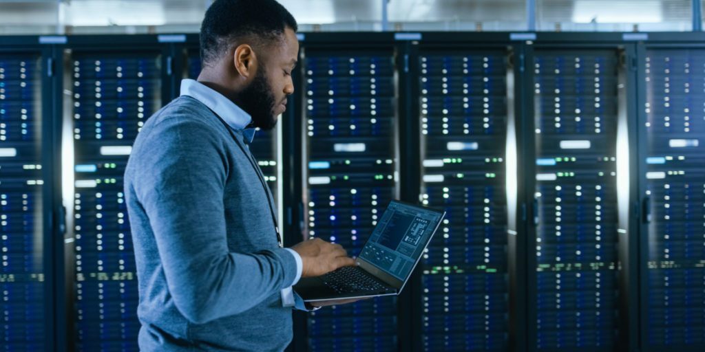 A man operates a laptop in a server room, surrounded by rows of servers with blue lights. He is deeply focused, embodying the expertise typical of IT consulting in North Jersey.