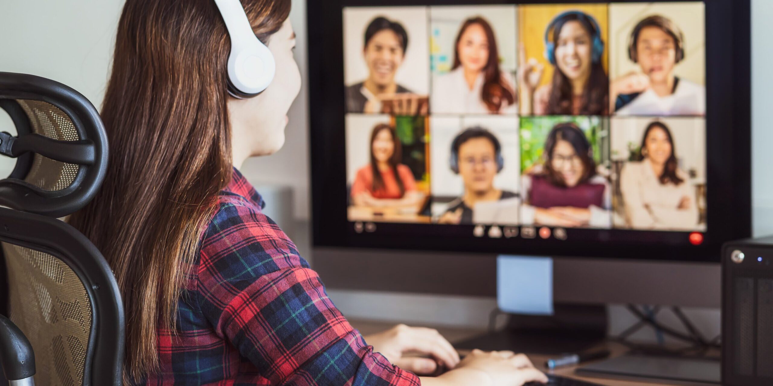 A person wearing headphones sits at a desk, participating in a video conference on Microsoft Teams. The screen displays a grid of eight diverse people, all appearing to be engaged in the virtual meeting. The person uses a keyboard and mouse to record Microsoft Teams meetings.