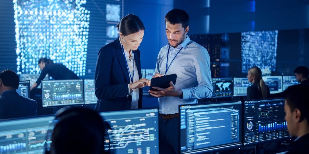 Two professionals review data on a tablet in a control room filled with multiple computer screens displaying various data charts and visuals, efficiently managing IT for banks.