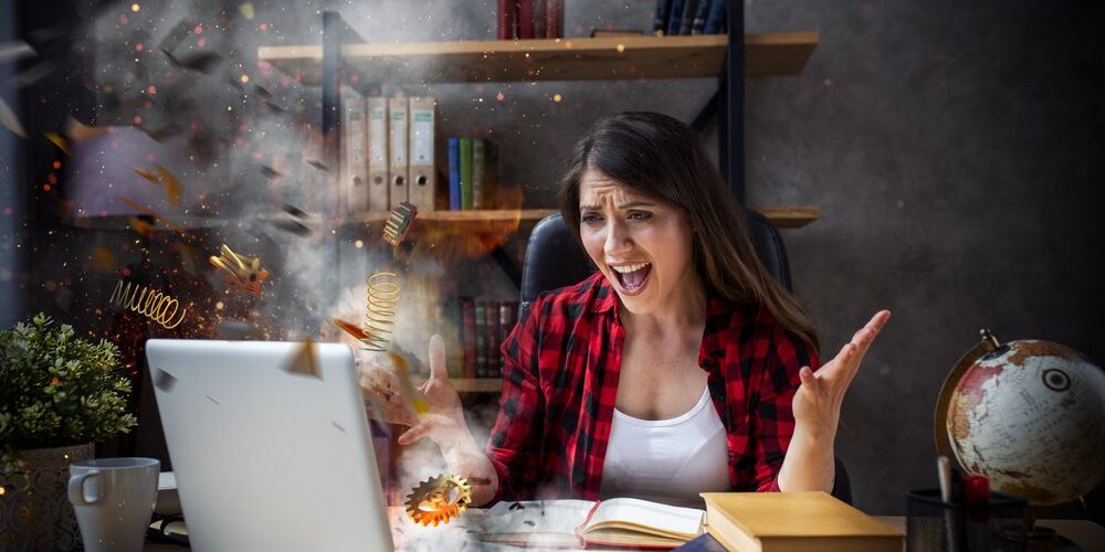 A woman wearing a red plaid shirt sits at a desk, looking frustrated as her laptop appears to be overheating, with springs and gears flying out. Her arms are raised in exasperation. The desk has books, a globe, a plant, and other office supplies on it.