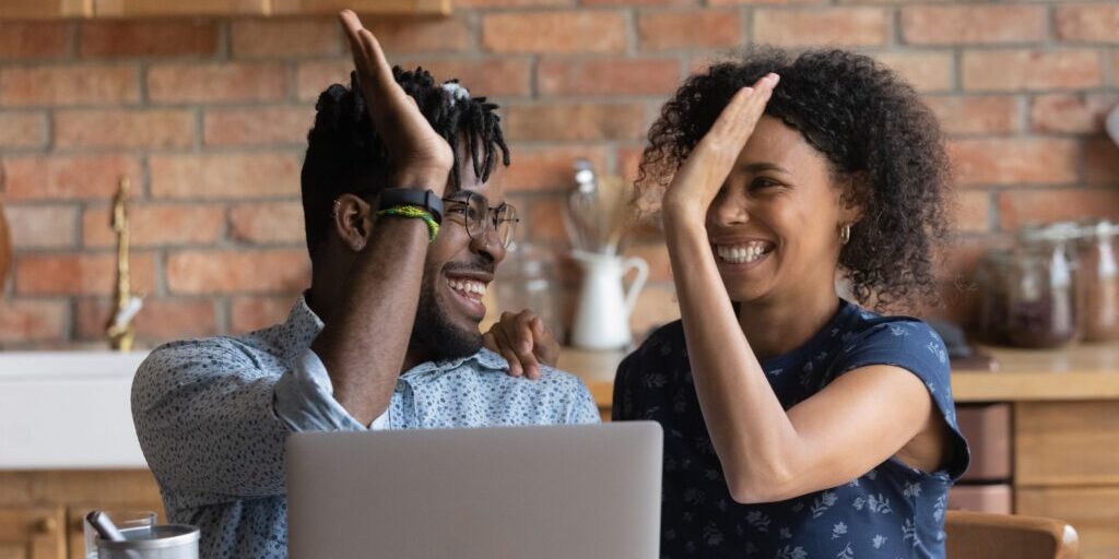 A smiling couple sits at a table in front of a laptop, giving each other a high five. The background features a brick wall and a cozy kitchen setting. The man wears glasses and a light blue shirt, while the woman is in a dark blue shirt with short sleeves.