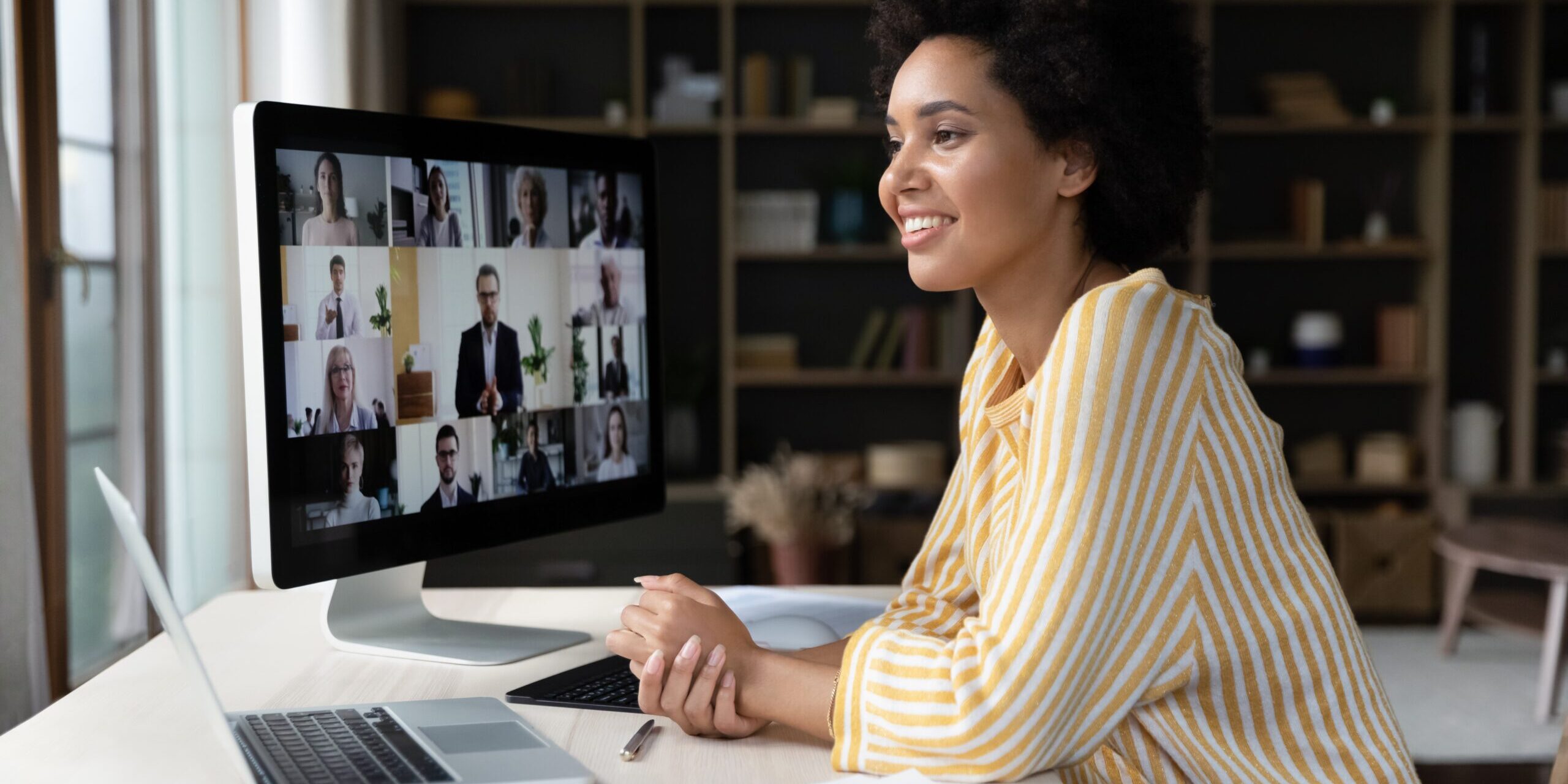 A woman in a yellow striped shirt smiles while attending a video conference on Microsoft Teams on her desktop computer. She is sitting at a desk with a laptop, papers, and a pen in a well-lit room with bookshelves in the background.