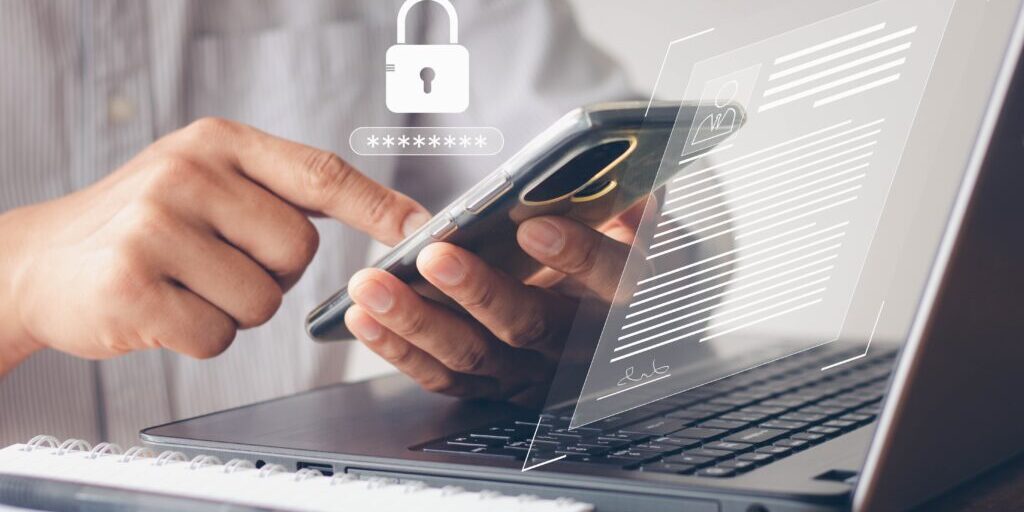 Person using a smartphone in front of a laptop with digital security icons, considering whether to change their password. A notebook and pen are placed on the table.