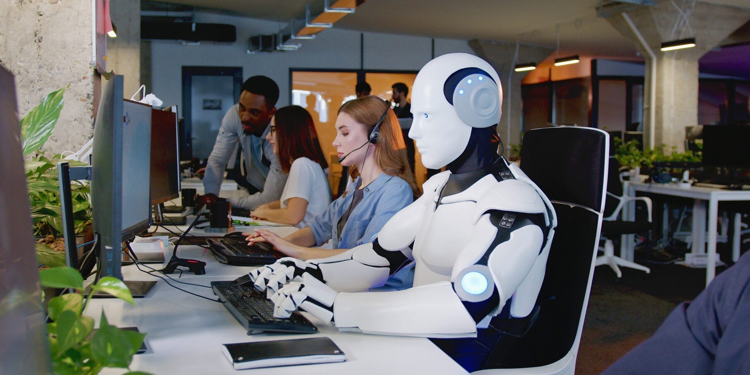 A humanoid robot and a woman wearing a headset work at computers in a modern office, guided by the AI Acceptable Use Policy. Other employees are in the background.