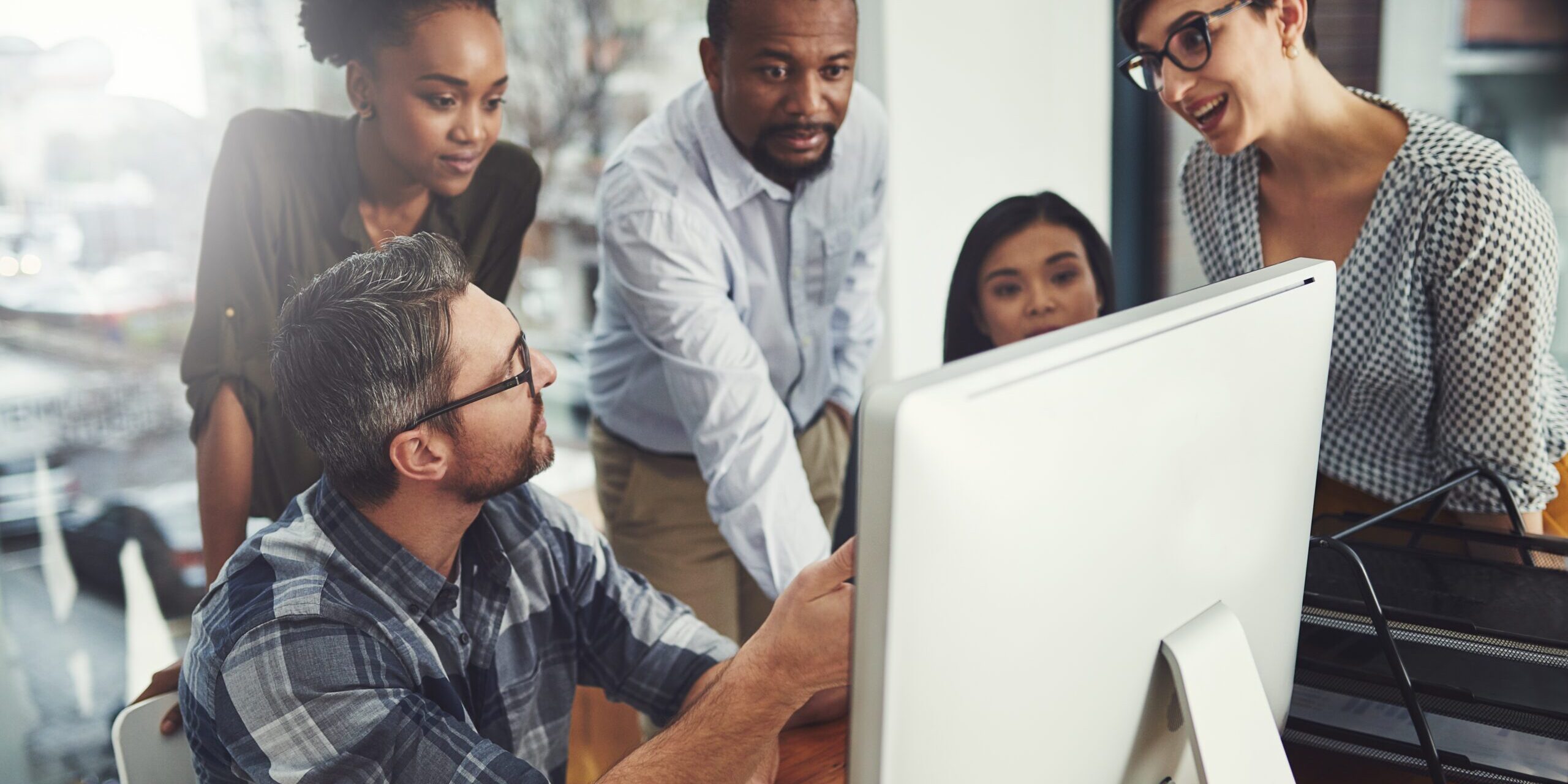 A group of five people is gathered around a computer in an office setting, exploring ways to customize Microsoft Teams. They appear engaged and are discussing something on the screen. The atmosphere looks collaborative and focused.
