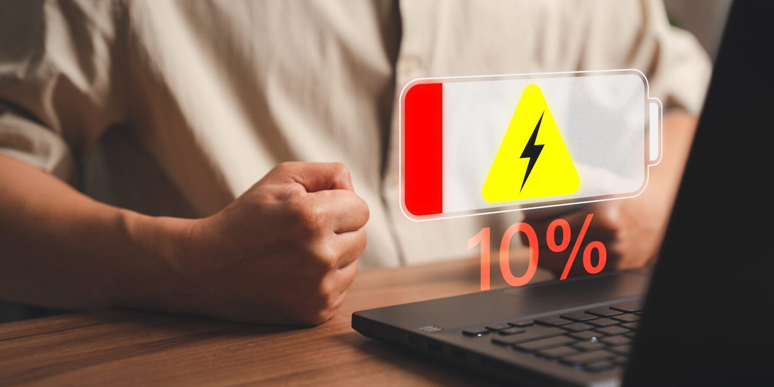 A person sits at a desk with a laptop. A low battery warning symbol, showing a lightning bolt and "10%" displayed in red, hovers above the screen. The individual's hand is clenched into a fist, possibly revealing frustration as the laptop battery drains rapidly.