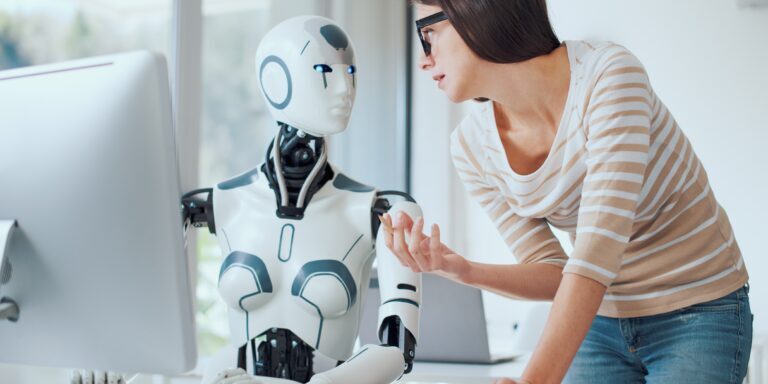 A woman in a striped shirt and glasses converses with a humanoid robot sitting at a computer desk, discussing the latest updates for Copilot for Microsoft Teams. The background features a modern office setting with large windows. The robot has a white body with black accents and blue lights for eyes.