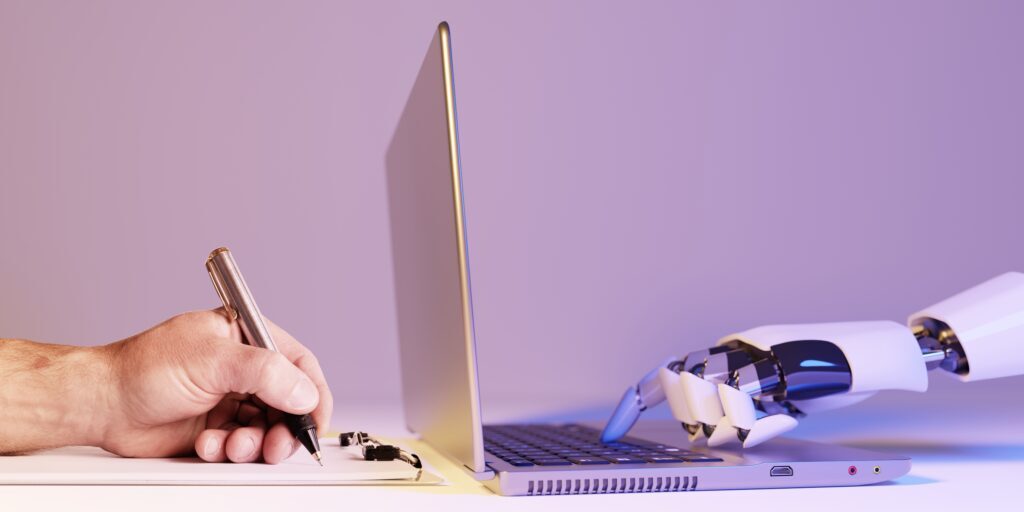 A human hand holding a pen writes on a notepad beside a laptop, while a robotic hand reaches out to the keyboard of the laptop. The scene emphasizes AI Readiness and highlighting the interaction between human and artificial intelligence.