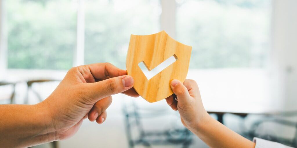 Two people holding together a wooden shield with a white checkmark, symbolizing protection or security. It's a visual representation of the reliability akin to cyber risk insurance. Blurred indoor background with natural light highlights their unity in safeguarding against threats.