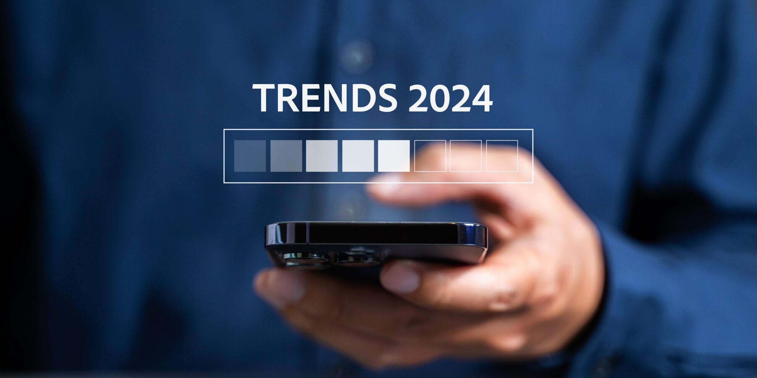 Closeup of a person in a blue shirt holding a smartphone, with "TRENDS 2024" and a horizontal loading bar displayed above the phone, indicating future trends and hot topics for cybersecurity in 2024. The background is blurred.