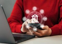 A person in a red shirt uses a smartphone in front of a laptop. Superimposed icons around the phone represent various digital banking and financial services, including a bank building, dollar sign, and bar chart—enhanced by AI for Banks.