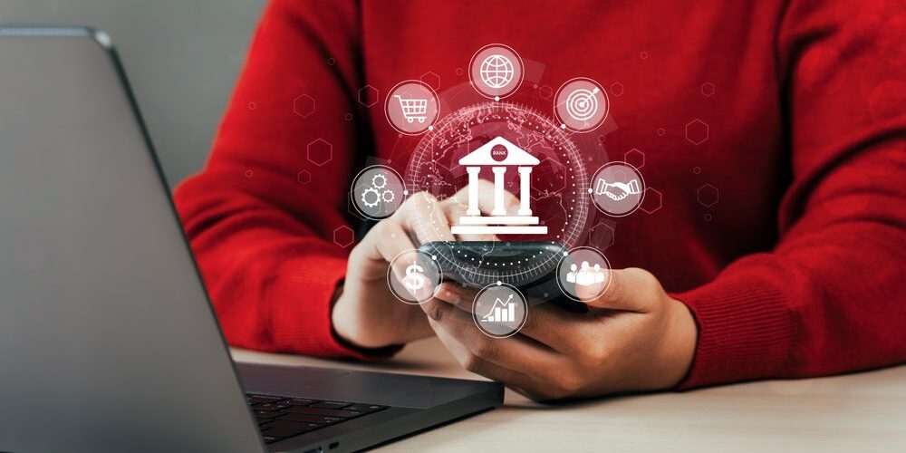 A person in a red shirt uses a smartphone in front of a laptop. Superimposed icons around the phone represent various digital banking and financial services, including a bank building, dollar sign, and bar chart—enhanced by AI for Banks.