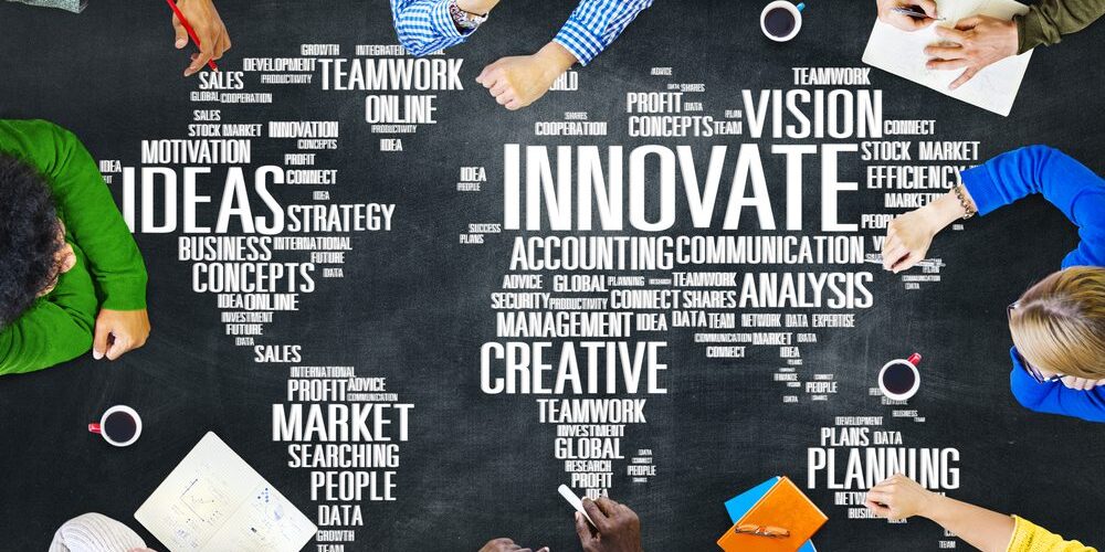 A group of people sit around a table covered with word clouds related to business, innovation, and teamwork, including terms like "ideas," "creative," "strategy," and "planning." Various documents and devices highlight their focus on developing an MSP for community banks.