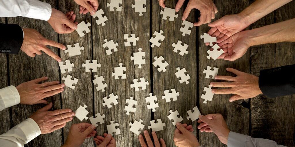 A group of people, whose hands are visible, are collaborating to assemble a jigsaw puzzle on a wooden table. The puzzle pieces, scattered around, are being picked up and arranged by various hands, indicating teamwork and cooperation. Much like the vCISO role in cybersecurity, everyone is pitching in to create the complete picture.