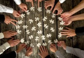 A group of people, whose hands are visible, are collaborating to assemble a jigsaw puzzle on a wooden table. The puzzle pieces, scattered around, are being picked up and arranged by various hands, indicating teamwork and cooperation. Much like the vCISO role in cybersecurity, everyone is pitching in to create the complete picture.
