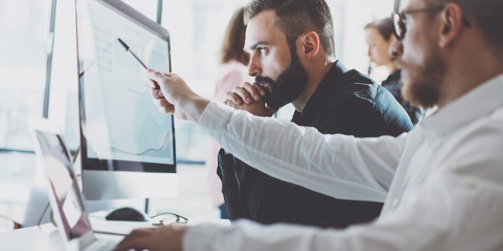 Two men at a desk working on a computer. One points at the screen displaying a graph while the other looks on thoughtfully.