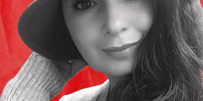 Black and white photo of a person with long hair wearing a hat, smiling against a red background.