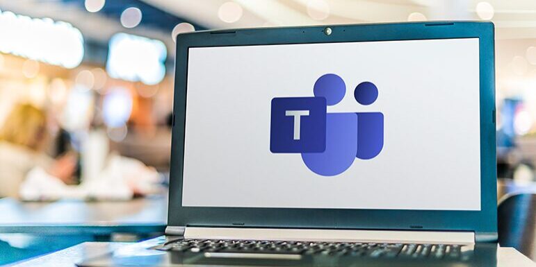 A laptop on a table with the Microsoft Teams logo displayed on the screen. The background is blurred, featuring people and lights.
