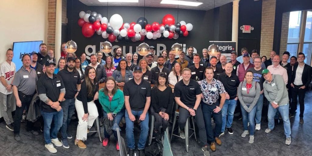 A large group of people posing for a photo in a room decorated with red, white, and black balloons. Most of them are standing, while a few are seated on stools in the front, celebrating the accomplishments of their IT support team in the Twin Cities.