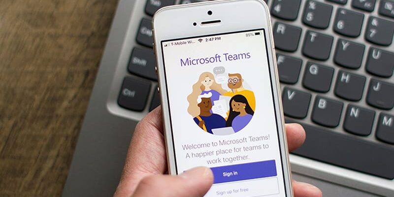 A person holding an iPhone displaying the "Microsoft Teams" welcome screen above a laptop keyboard.