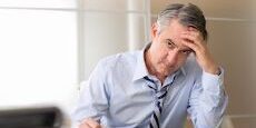 A man in a shirt and tie sits at a desk with his hand on his forehead, appearing stressed or tired. Papers and a pen are on the desk.